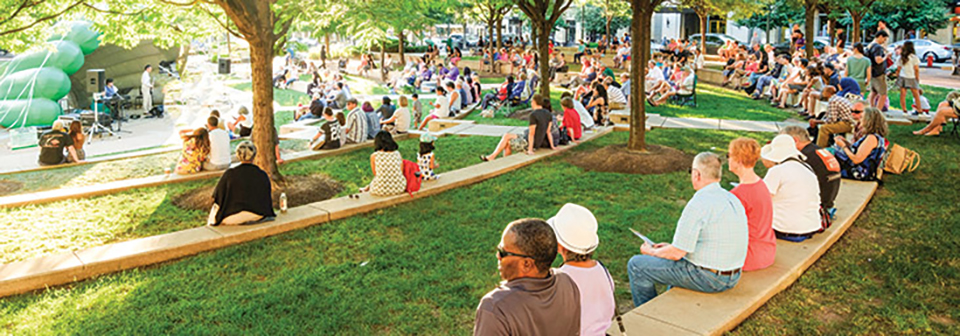 People outside seated on benches in a grassy park listening to an outdoor concert.