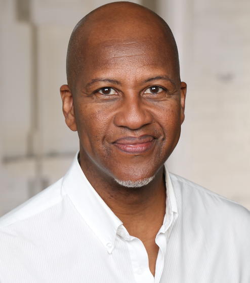 Professional headshot of Djola Branner, wearing a white shirt and smiling into the camera